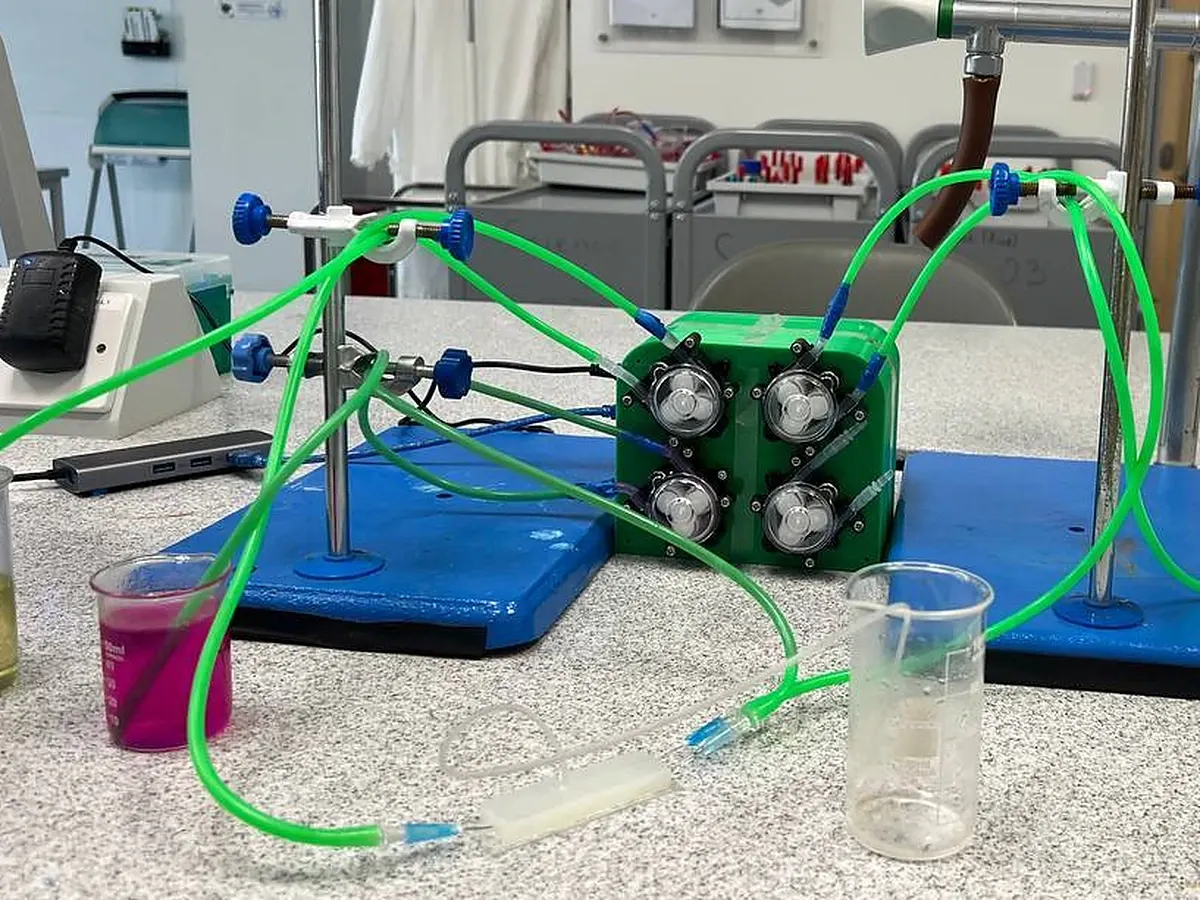 The four-channel peristaltic pump in its green 3D-printed housing, tubing running to a PDMS chip and beakers on a school lab bench